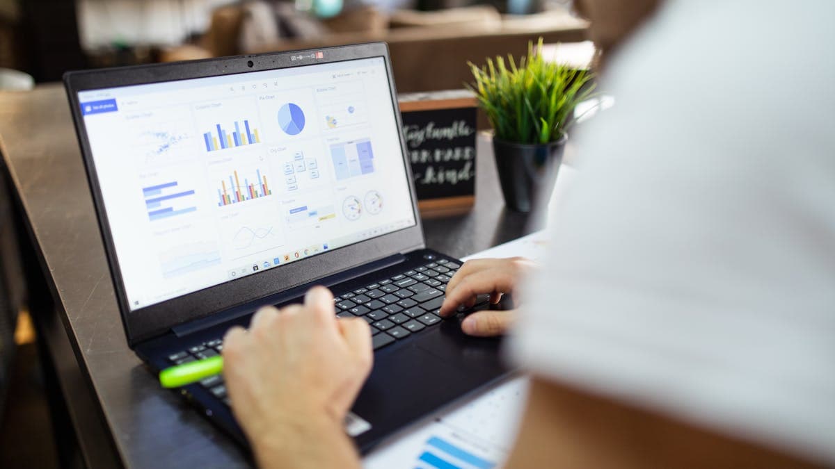 A cluttered desk with a laptop showing a spreadsheet and stacks of paperwork